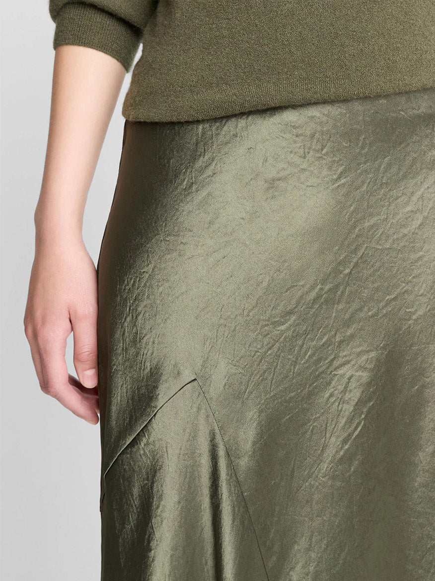 A close-up of a person wearing an olive green sweater and the Vince Paneled Satin Bias Skirt in Herba, featuring shiny, slightly wrinkled Naia acetate satin with an asymmetric design.