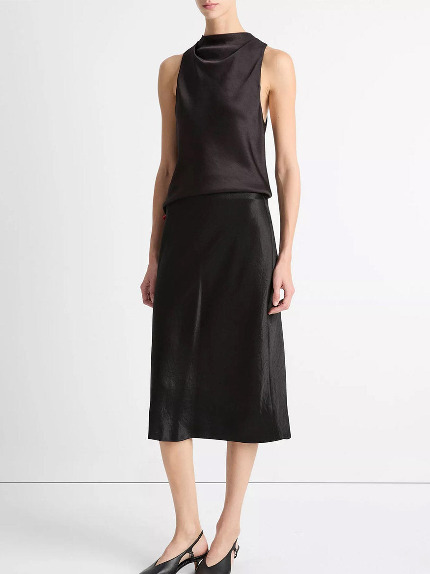 A person wearing a sleeveless black draped top, the Vince Satin Slip Skirt in Black, and black slingback shoes stands against a plain white background.