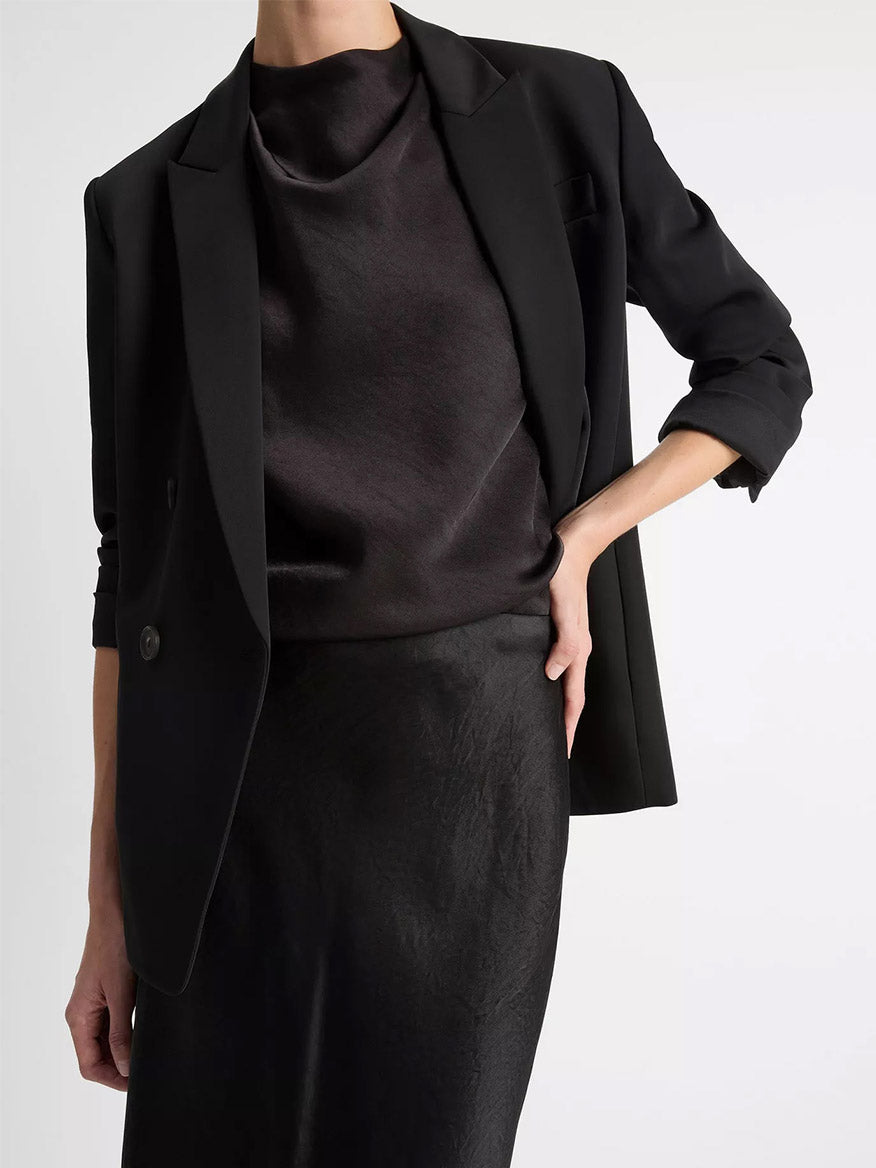 A person stands against a plain background with one hand on hip, wearing a black blazer over the Vince Satin Slip Skirt in Black and a matching top.