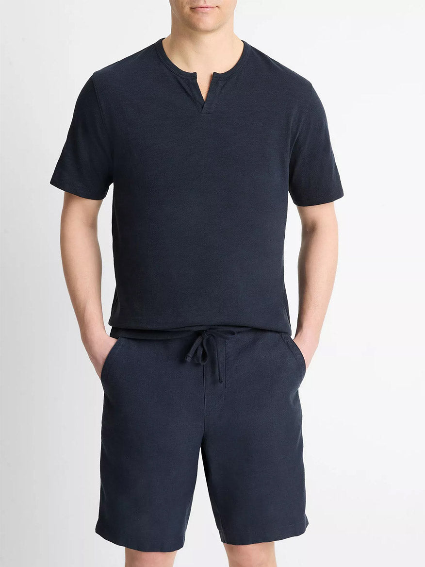 A man stands against a plain background with his hands in his pockets, wearing the Vince Slub Cotton Split-Neck T-Shirt in Coastal Blue and matching drawstring shorts.