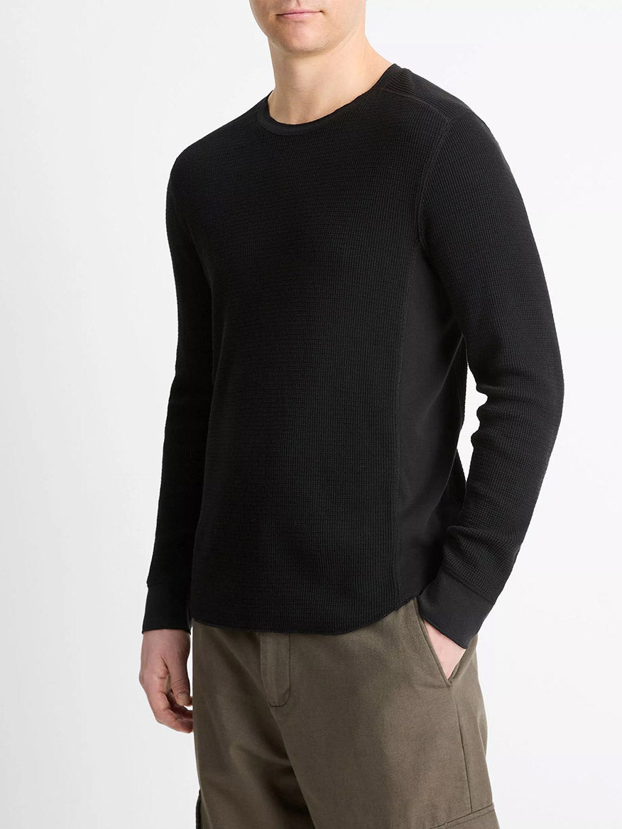 A man wears the Vince Pima Cotton-Blend Thermal Long-Sleeve Pullover in black with olive-green pants, standing with one hand in his pocket against a plain white background.