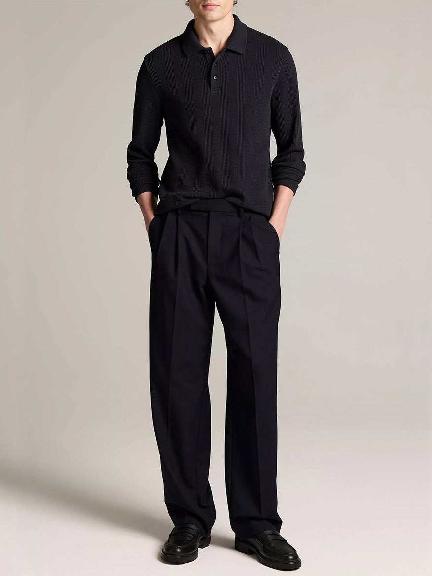 A man stands against a plain background in the Vince Variegated Cotton Polo Shirt in Coastal Blue, dark pleated trousers, and black shoes, hands in pockets, face partially out of frame.