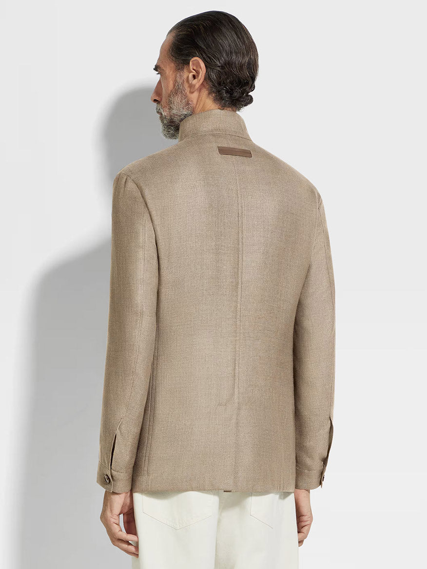 A man with slicked-back hair, wearing a Zegna Il Conte Chore Jacket in Light Taupe and white trousers, stands with his back to the camera against a plain, light background.