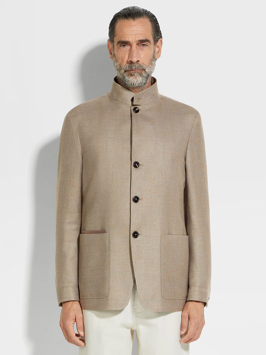 A man with gray hair and beard wears a Zegna Il Conte Chore Jacket in Light Taupe, featuring dark buttons and large front pockets, paired with white trousers as he stands against a plain background.