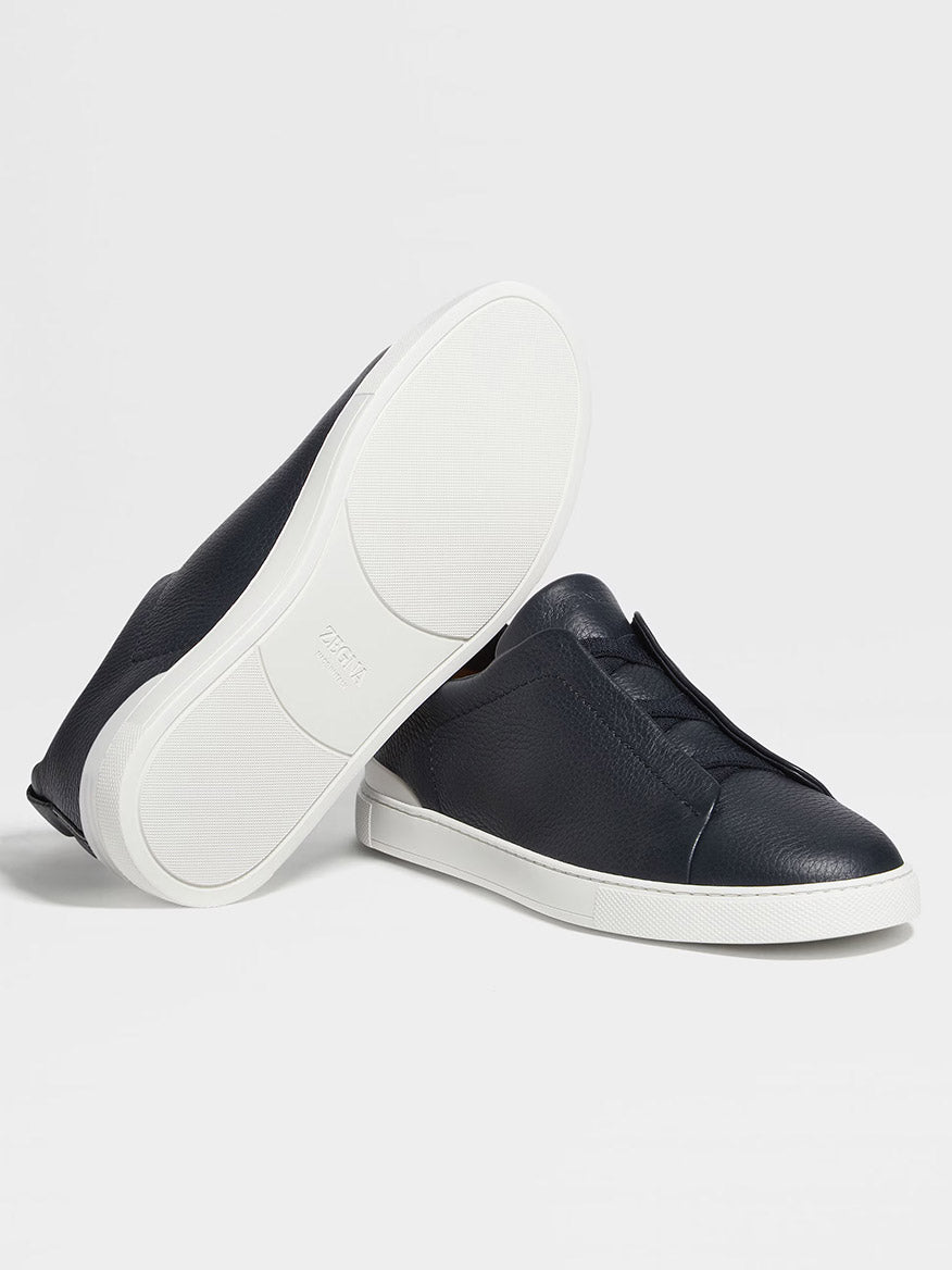 A pair of navy blue Zegna Triple Stitch™ sneakers in premium deerskin, featuring a slip-on low-top design with white rubber soles—one upright, the other showing the sole.