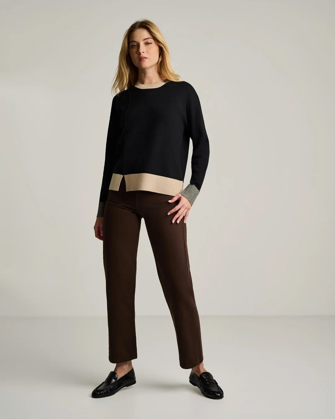 A woman stands in a neutral room wearing the Lisa Todd Bold Block Cotton Cashmere Sweater in Black, paired with brown pants and black shoes.