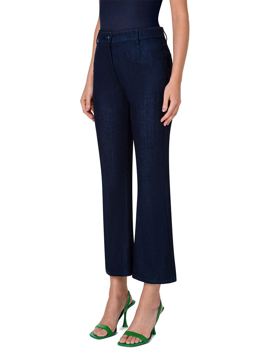 A person wearing Akris Punto Cali Stretch Crop Bootcut Denim in Dark Blue, a black top, and green high-heeled sandals stands against a plain background.