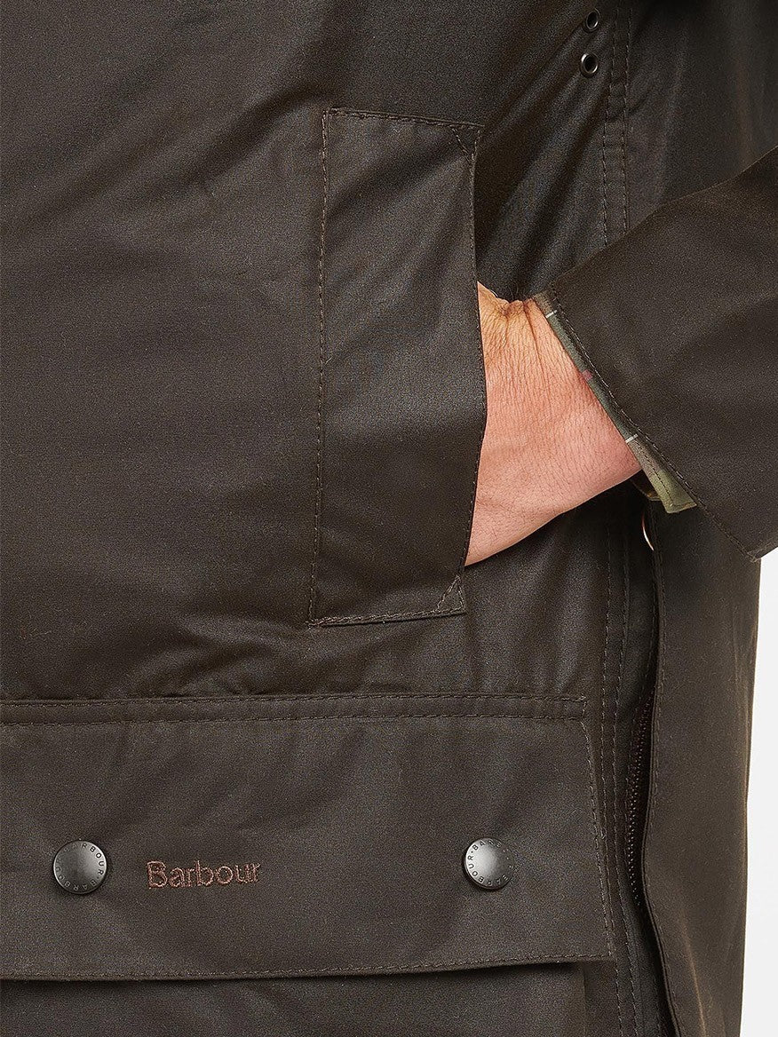 Close-up of a person wearing a dark Barbour Classic Beaufort in Olive with a quilted liner, their hand in the pocket showcasing the brand logo and button details.