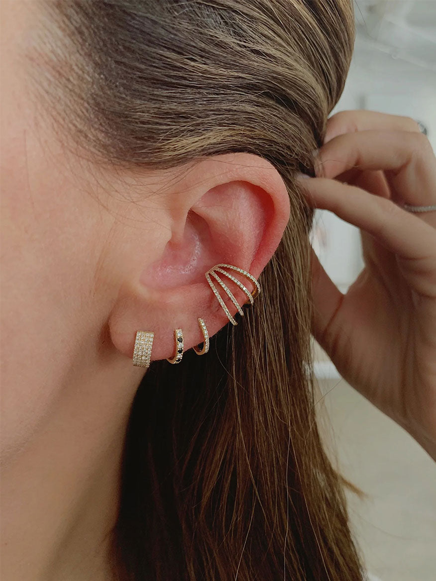 Close-up of a person’s ear adorned with multiple earrings including a diamond stud, the EF Collection Diamond Huggie Earrings in Yellow Gold, a triple-chevron design, and a spiral cuff. Hand pulling back hair to better reveal the jewelry.