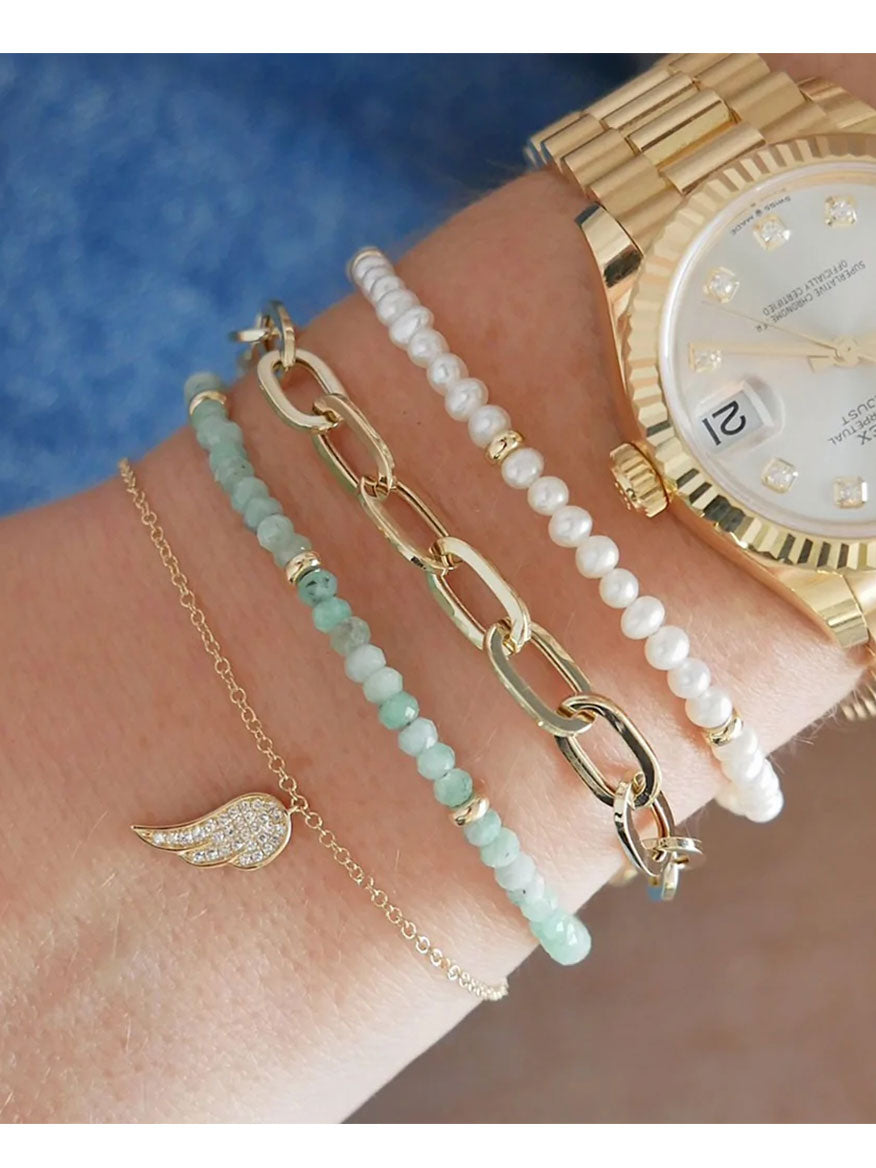 Close-up of a wrist adorned with a 14k gold watch and four bracelets: EF Collection Birthstone Bracelet with Gold Rondelles - Pearl, green beads, gold links, and a delicate charm bracelet featuring natural stones and a small wing.