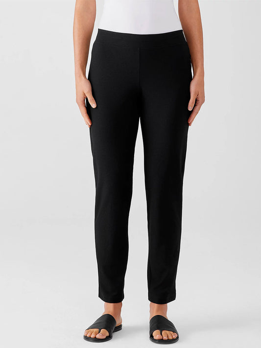 A person wearing slim Eileen Fisher Washable Stretch Crepe Pants in Black and sandals stands against a neutral background.