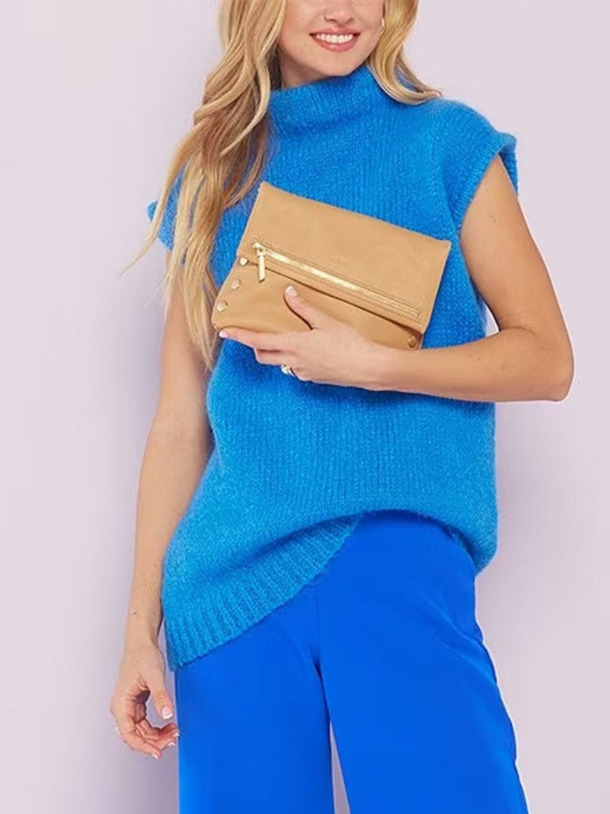 Woman posing with a blue sleeveless turtleneck and matching pants, holding a beige Hammitt Los Angeles VIP Medium Clutch in Toast Tan.