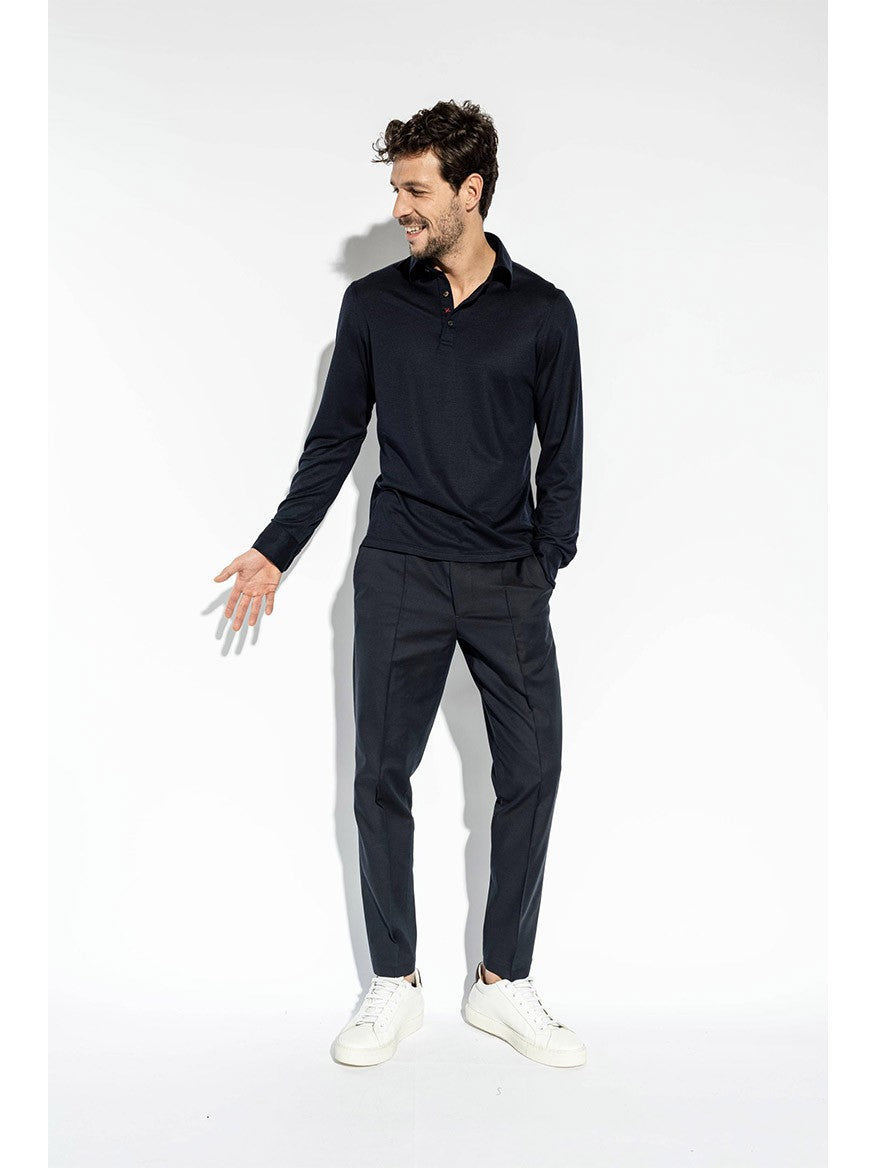 A man wearing an Isaia Evening Polo in Navy, dark pants, and white sneakers stands against a white background, looking to his left with a slight smile.