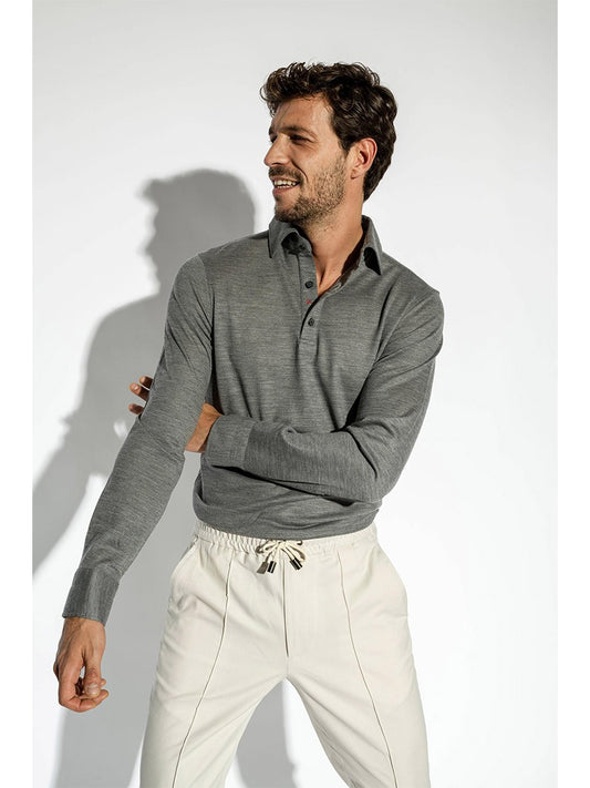 A man in a gray long-sleeve Isaia Evening Polo shirt in Medium Grey and white drawstring pants stands smiling with his arms crossed, casting a shadow on a plain wall.