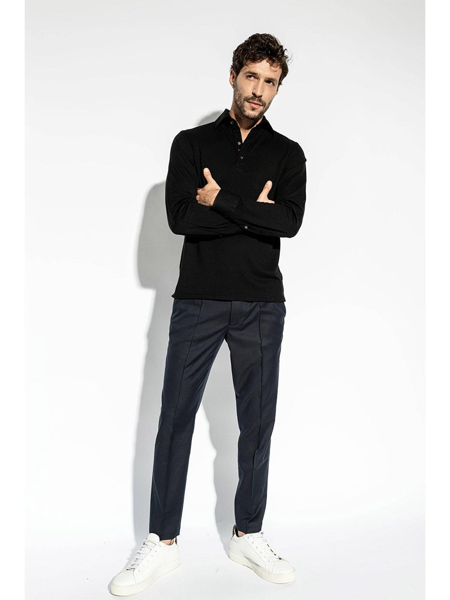 A man with dark curly hair wearing an Isaia Evening Polo in Black, blue pants, and white sneakers stands against a plain white background.