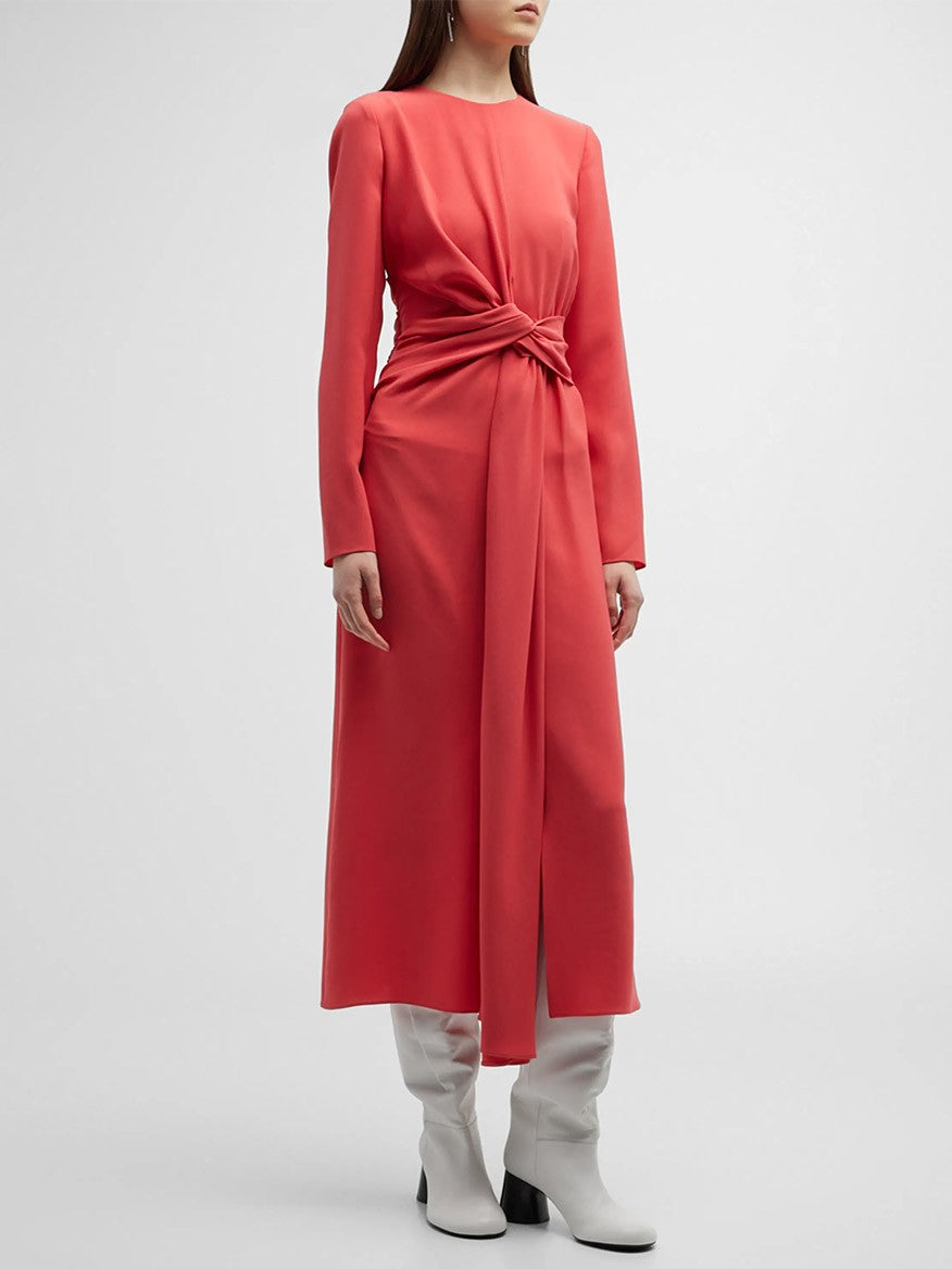 A person stands against a plain background, wearing a Lafayette 148 New York Finesse Crepe Twist Dress in Coral, featuring a front twist at the waist and white ankle boots. The A-line silhouette flatters the figure, though their face remains out of view.