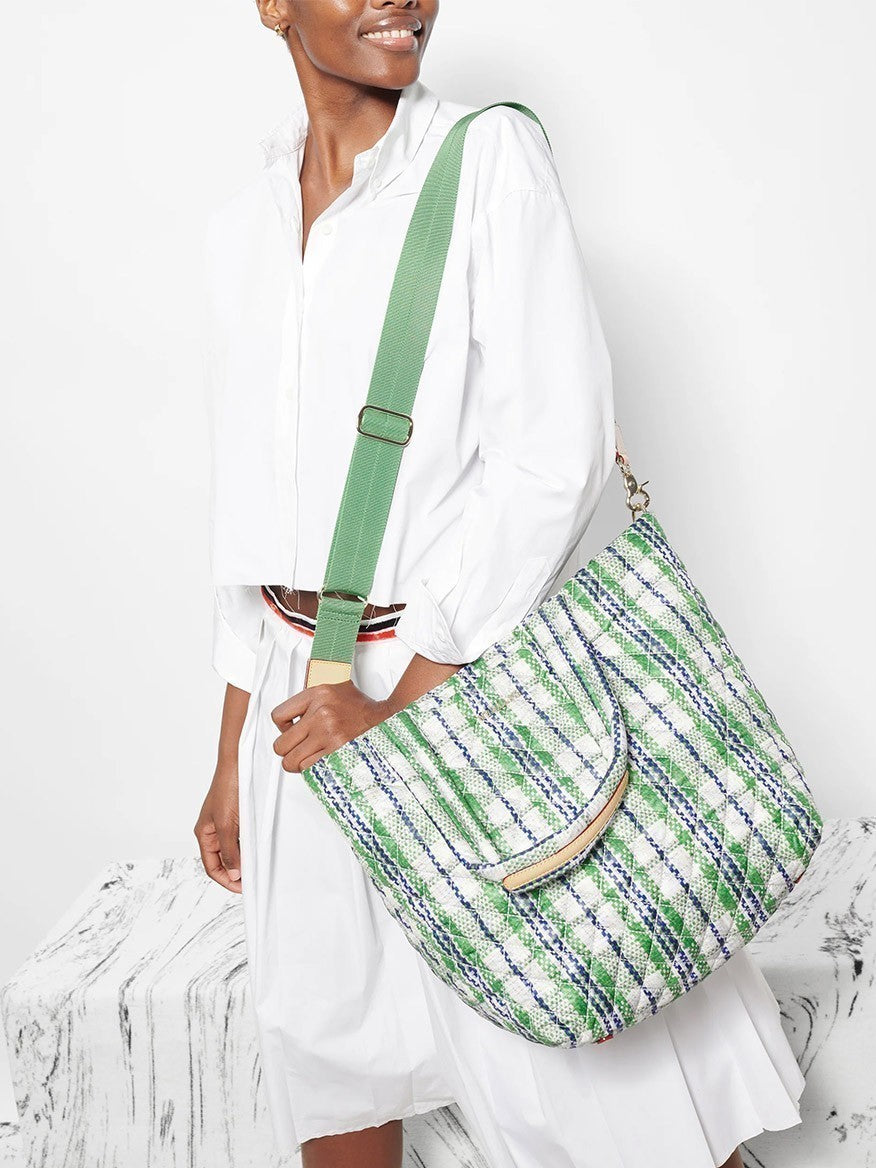 A woman in a white dress carries a large green striped MZ Wallace Medium Metro Tote Deluxe in Spring Plaid Oxford with a brown handle, standing against a white background.