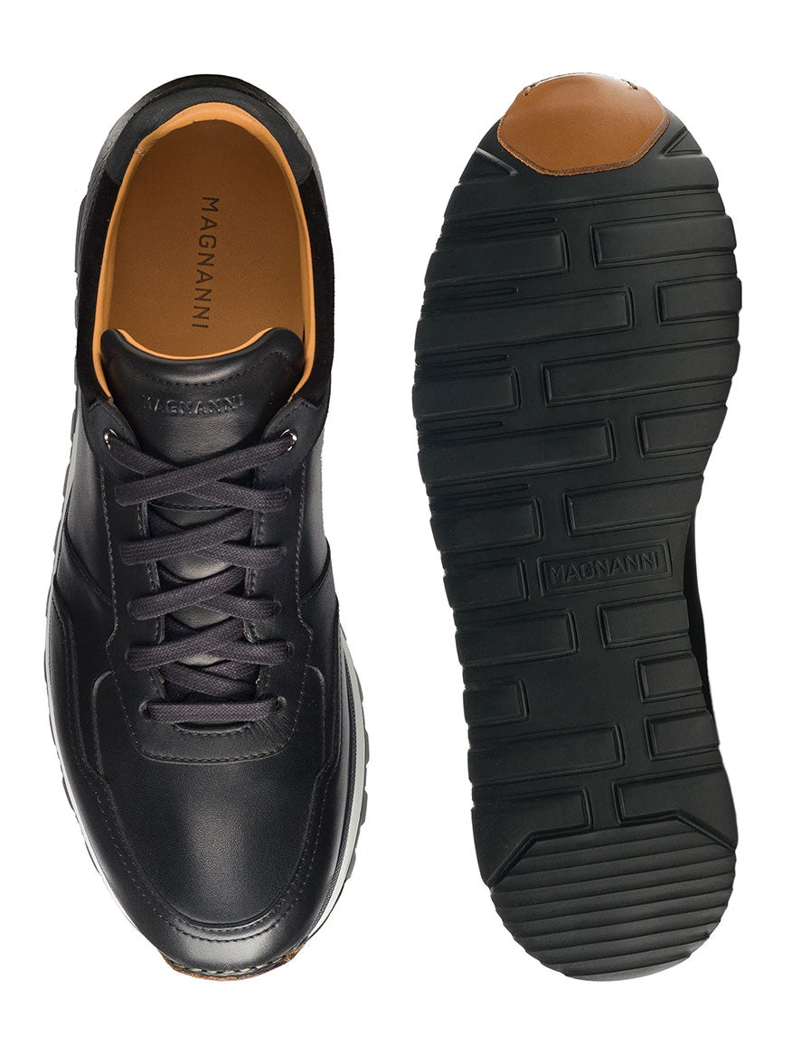 Magnanni Arco in Black showcases black leather sneakers with rubber soles; one shoe from the top highlights hand-stitched leather toe and laces, while the other reveals a tread pattern. Retro-inspired design with brown interior accents ensures a lightweight and flexible feel.