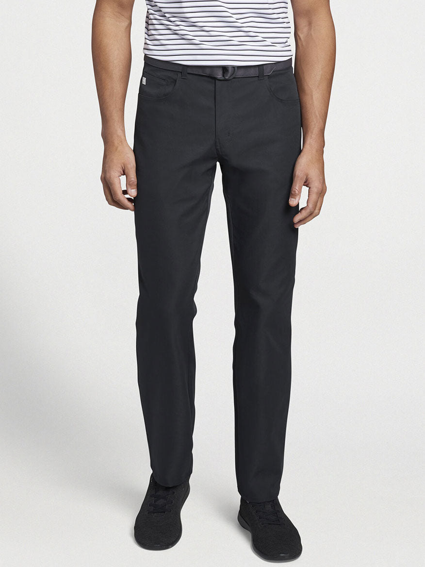 A person wearing a black and white striped shirt, Peter Millar Performance Five-Pocket Pant in Black with two-way stretch, a belt, and sleek black shoes.