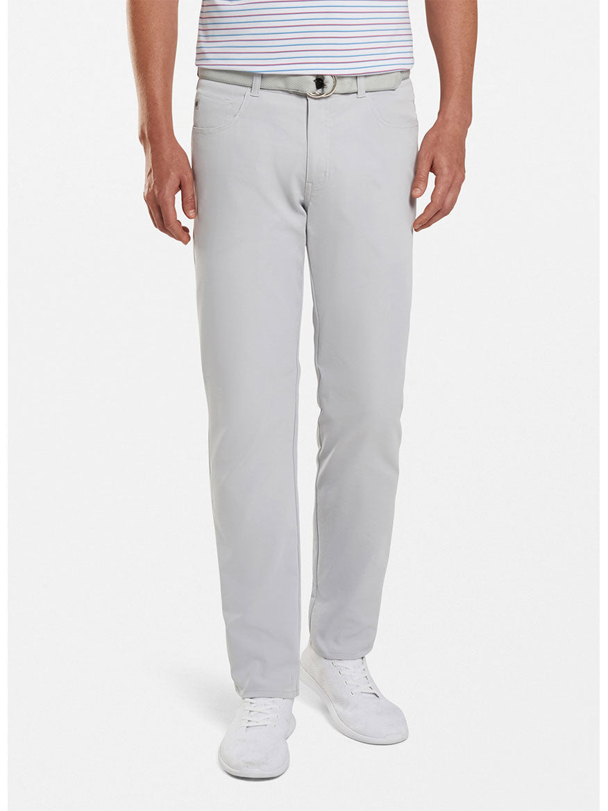 A man wearing Peter Millar Performance Five-Pocket Pants in British Grey made of quick-drying fabric, a white belt, a striped shirt, and white shoes stands against a white background.