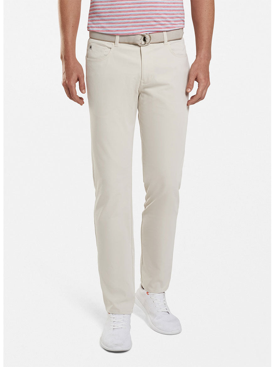 Man wearing Peter Millar Performance Five-Pocket Pant in Stone, a striped t-shirt, and white sneakers, standing with hands in pockets against a white background.