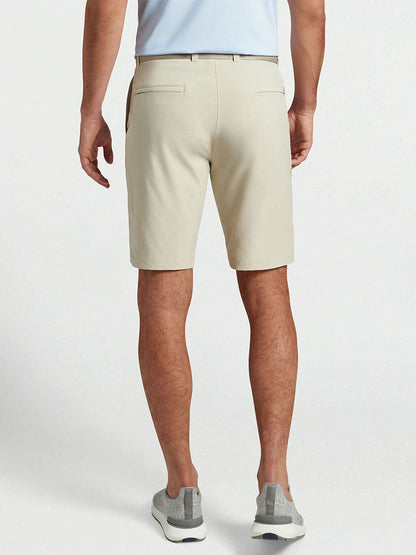 The back view of a man wearing the Peter Millar Shackleford Performance Hybrid Short in Sand and a beige shirt, enjoying sunny days on the course.