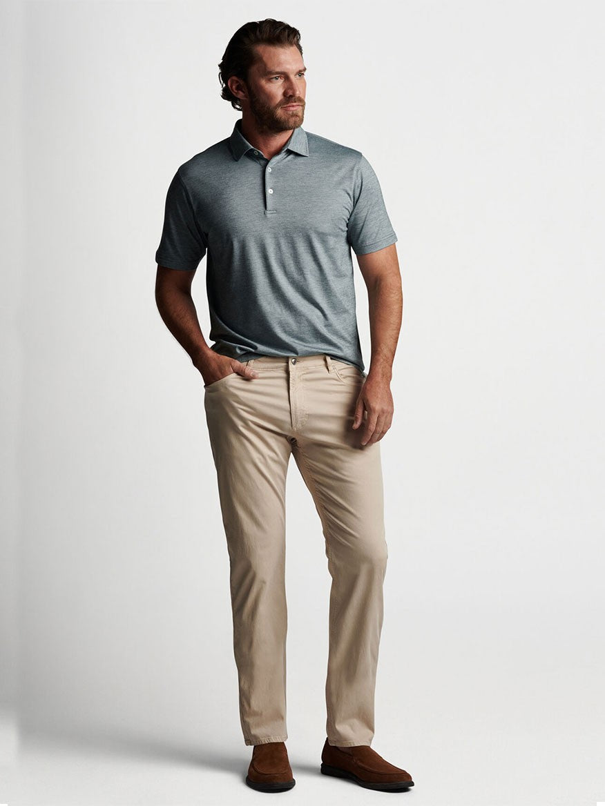 A man in a tailored fit grey polo shirt and beige Peter Millar Wayfare Five-Pocket Pant in Stone posing against a white background.
