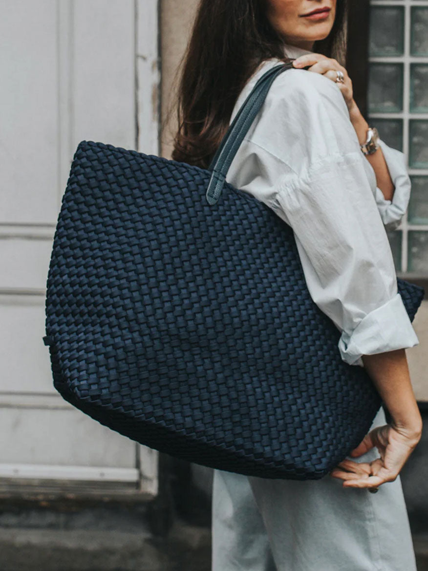 A person in a white shirt and gray pants carries the spacious Naghedi Jetsetter Large Tote in Solid Ink Blue over their shoulder, ideal for travel.