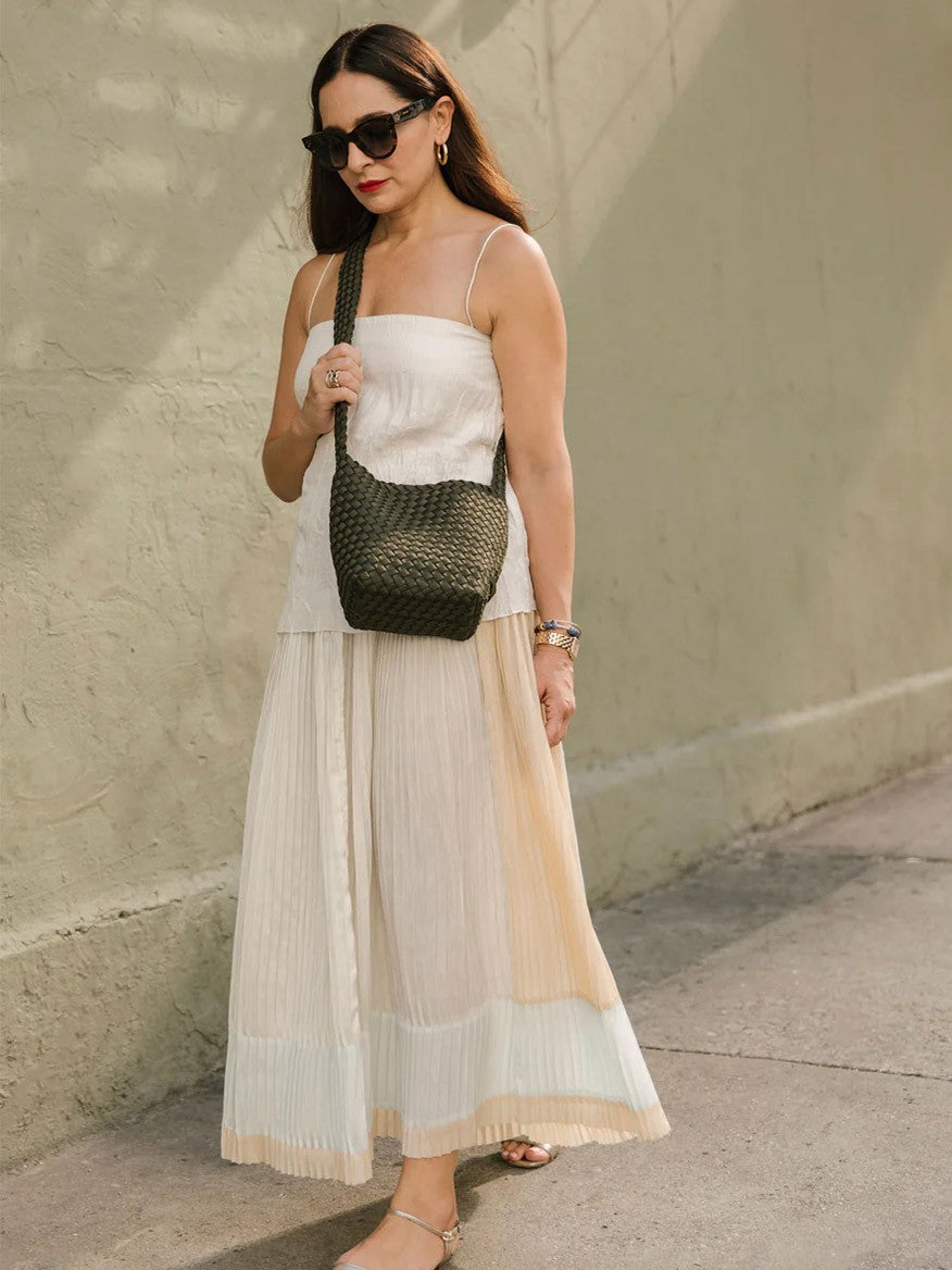 Woman in a white dress with a pleated skirt and sunglasses carries a Naghedi Laguna Crossbody in Solid Onyx while standing against a beige wall in sunlight.