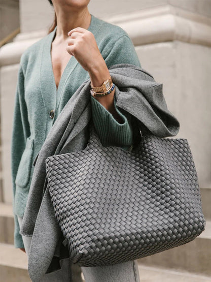 A woman in a teal cardigan carrying a Naghedi St. Barths Medium Tote in Solid Onyx and a draped gray coat over her arm.