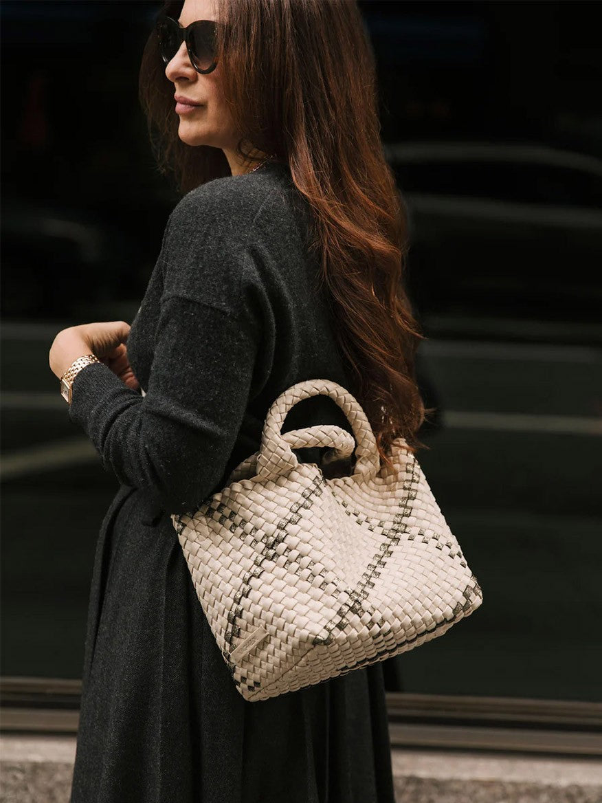 A woman with long brown hair wearing a dark coat holds a chic and compact Naghedi St. Barths Small Tote in Solid Onyx. She stands near a window with a dark reflection, wearing sunglasses.