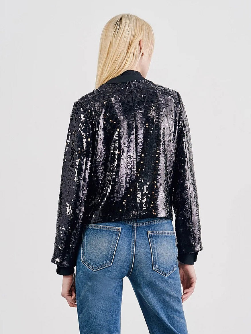 A person with long blonde hair is shown from the back, wearing a Nili Lotan Katherine Sequin Jacket in Black adorned with embroidered gold buttons and blue jeans.