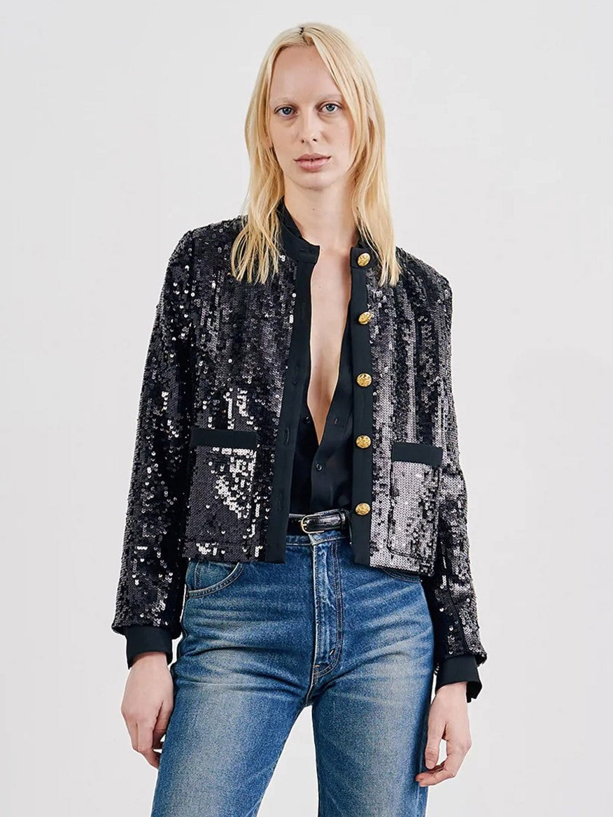 A person with long blonde hair is wearing a black Nili Lotan Katherine Sequin Jacket adorned with embroidered gold buttons, a dark shirt, and blue jeans, standing against a plain background.