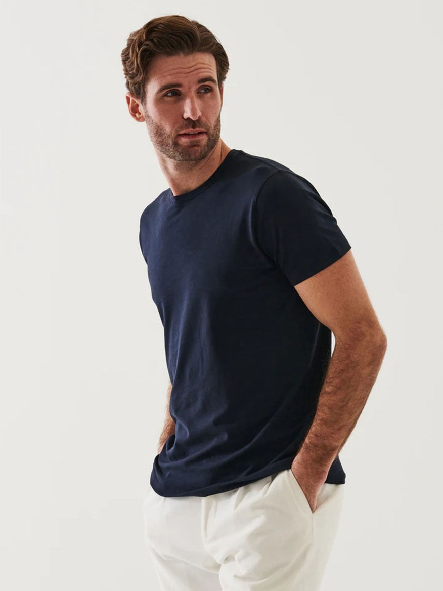 A man with short brown hair, beard, and mustache, looking to the side, wearing a dark blue Patrick Assaraf Iconic T-Shirt in Midnight and white pants, standing against a plain white background.