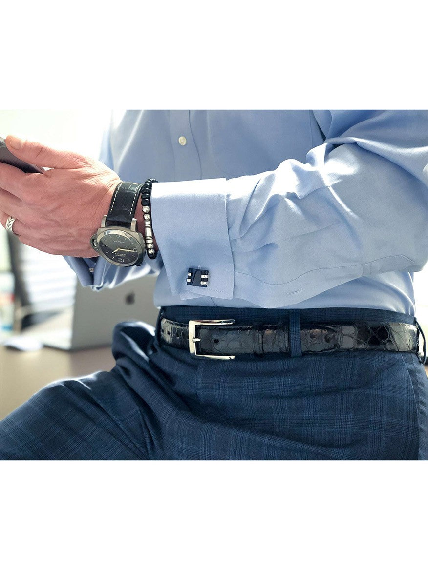 A person in a blue dress shirt and plaid trousers, wearing a wristwatch and bracelets, is holding a smartphone. A handmade Torino Leather American Alligator Skin Belt in Black with a polished nickel buckle is also visible.