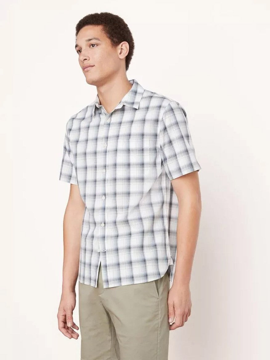 A man stands wearing a Vince Atwater Plaid Short Sleeve Shirt in Celery, made of pure cotton with a classic fit and light-gray and white plaid pattern, paired with beige pants, looking off to the side.