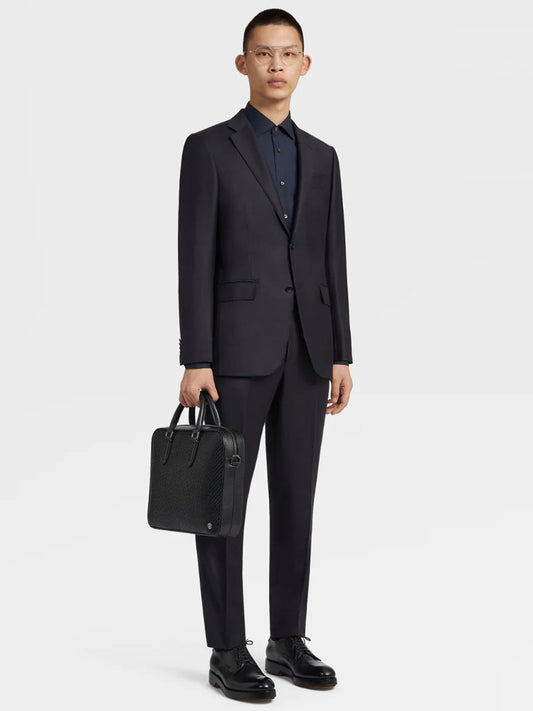 A person in a Zegna Navy Blue Milano Trofeo™ Wool Suit with a navy blue button-up shirt holds a black leather briefcase. They stand against a plain white background.