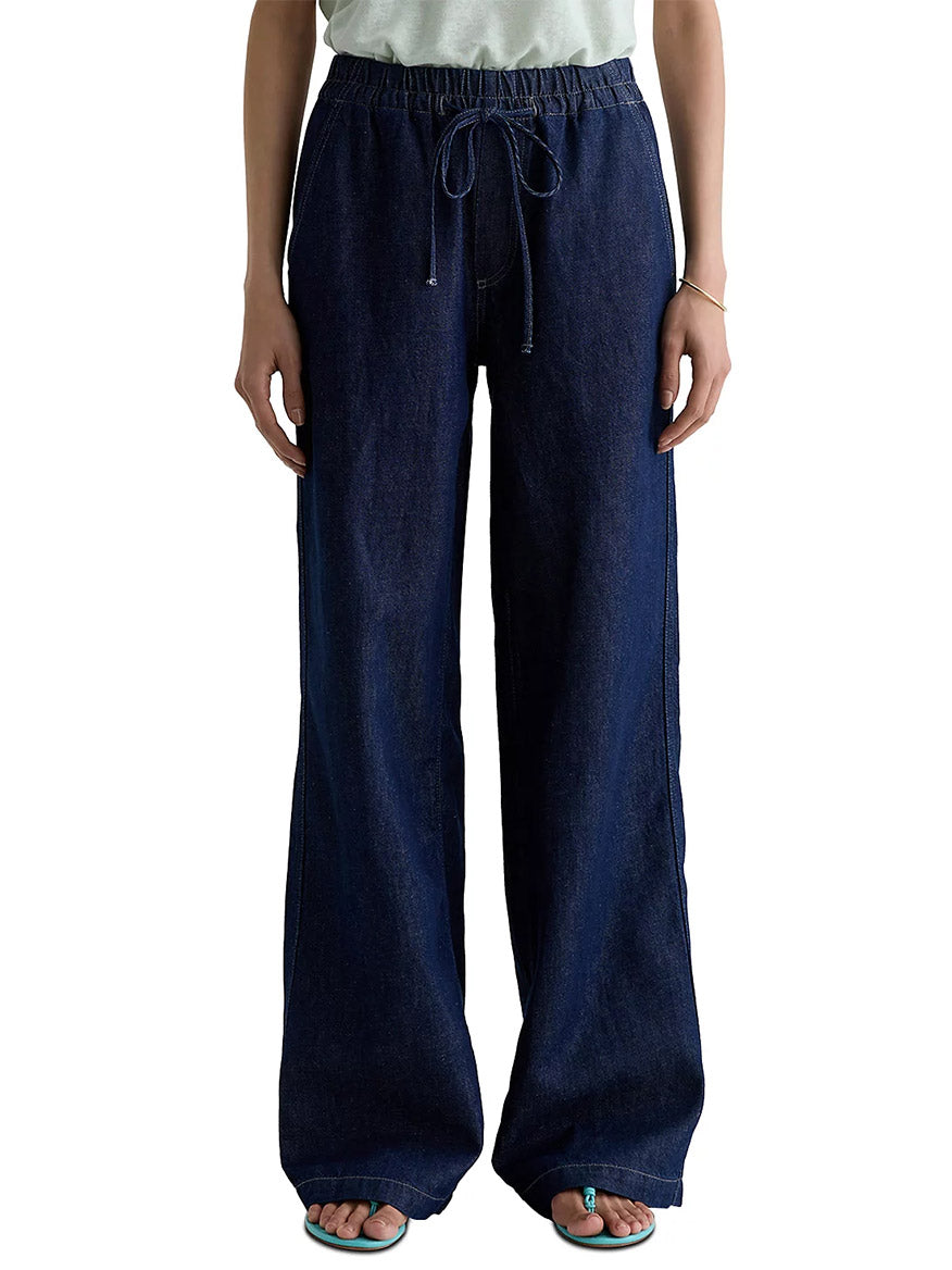 A person wearing AG Jeans Adria Cinched Jean Low-Rise Wide Leg in Napa, featuring a loose fit, dark blue linen-blend denim, elastic waistband, and drawstring, stands with hands by their sides.