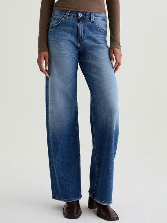 Person wearing AG Jeans Adria Low-Rise Wide Leg in Headline, premium blue Japanese denim jeans, and a brown long-sleeve top, standing against a plain light background.