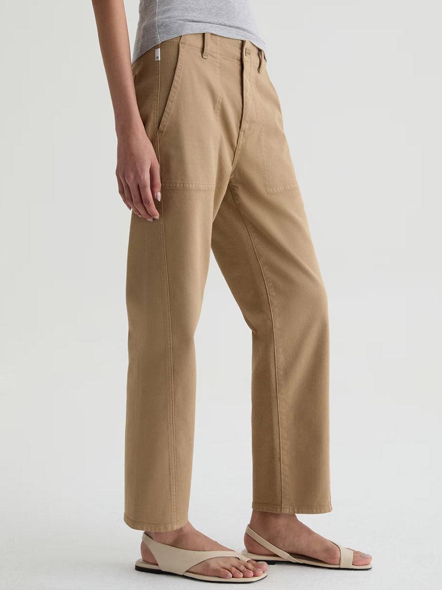 Model wears AG Jeans Analeigh Mid-Rise Utility Pant in Khaki Sand, paired with a light gray top and beige sandals, standing against a plain white background.