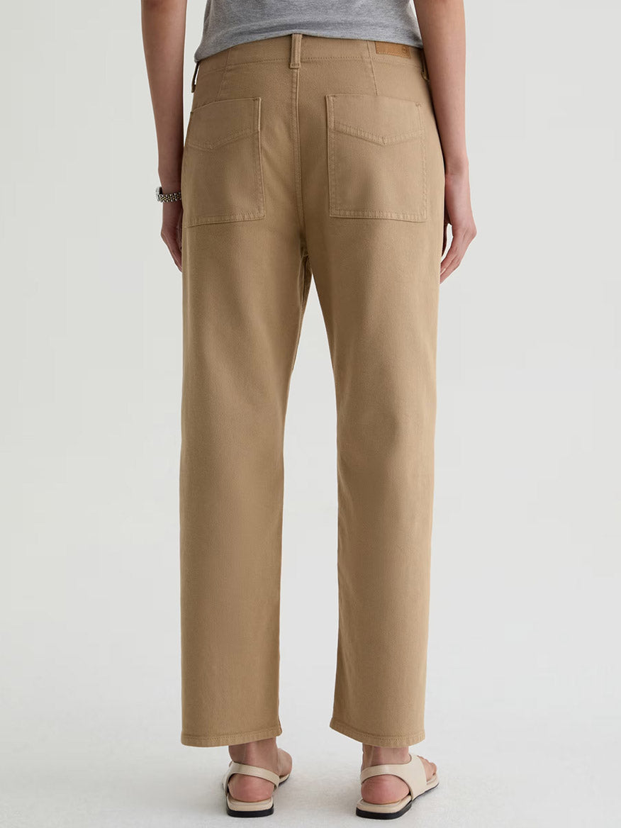 A person stands facing away, wearing AG Jeans Analeigh Mid-Rise Utility Pant in Khaki Sand, a gray shirt, sandals, and a bracelet against a plain light background.