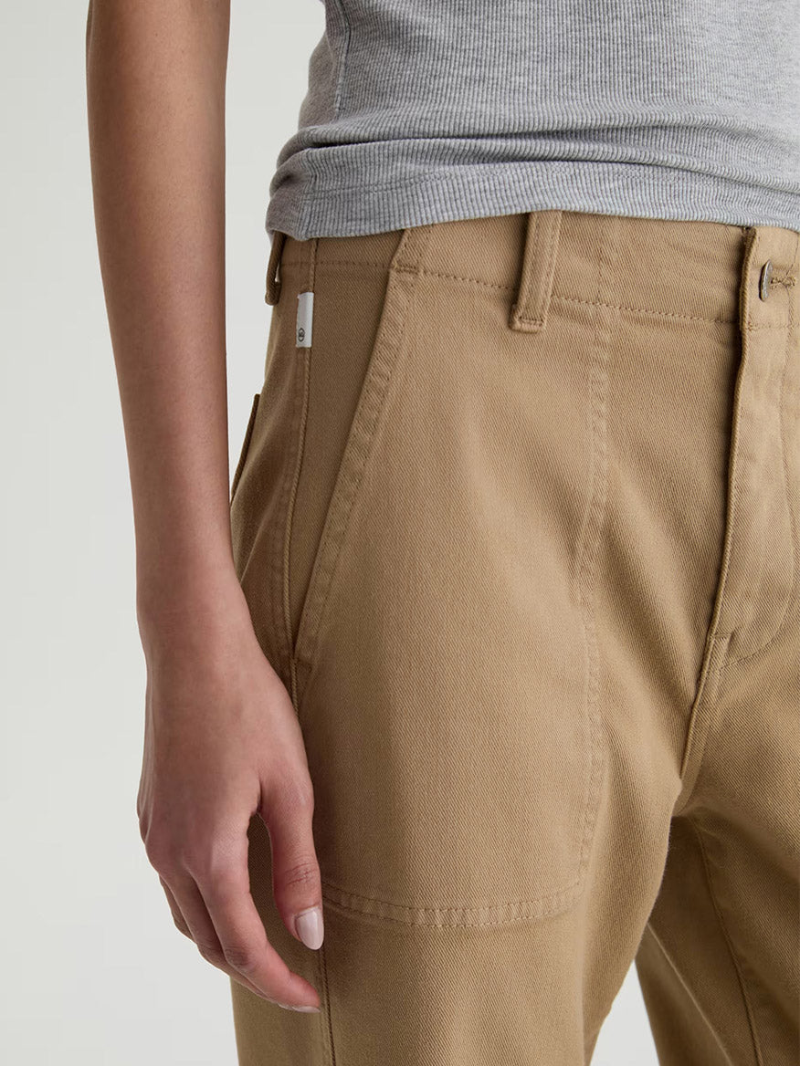 A person shown from the waist to mid-thigh wears a gray sleeveless top paired with AG Jeans Analeigh Mid-Rise Utility Pant in Khaki Sand, featuring front pockets.