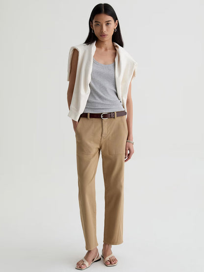 A woman faces forward in a grey tank top, the AG Jeans Analeigh Mid-Rise Utility Pant in Khaki Sand with a brown belt, a white sweater over her shoulders, and sandals. The background is plain and light-colored.