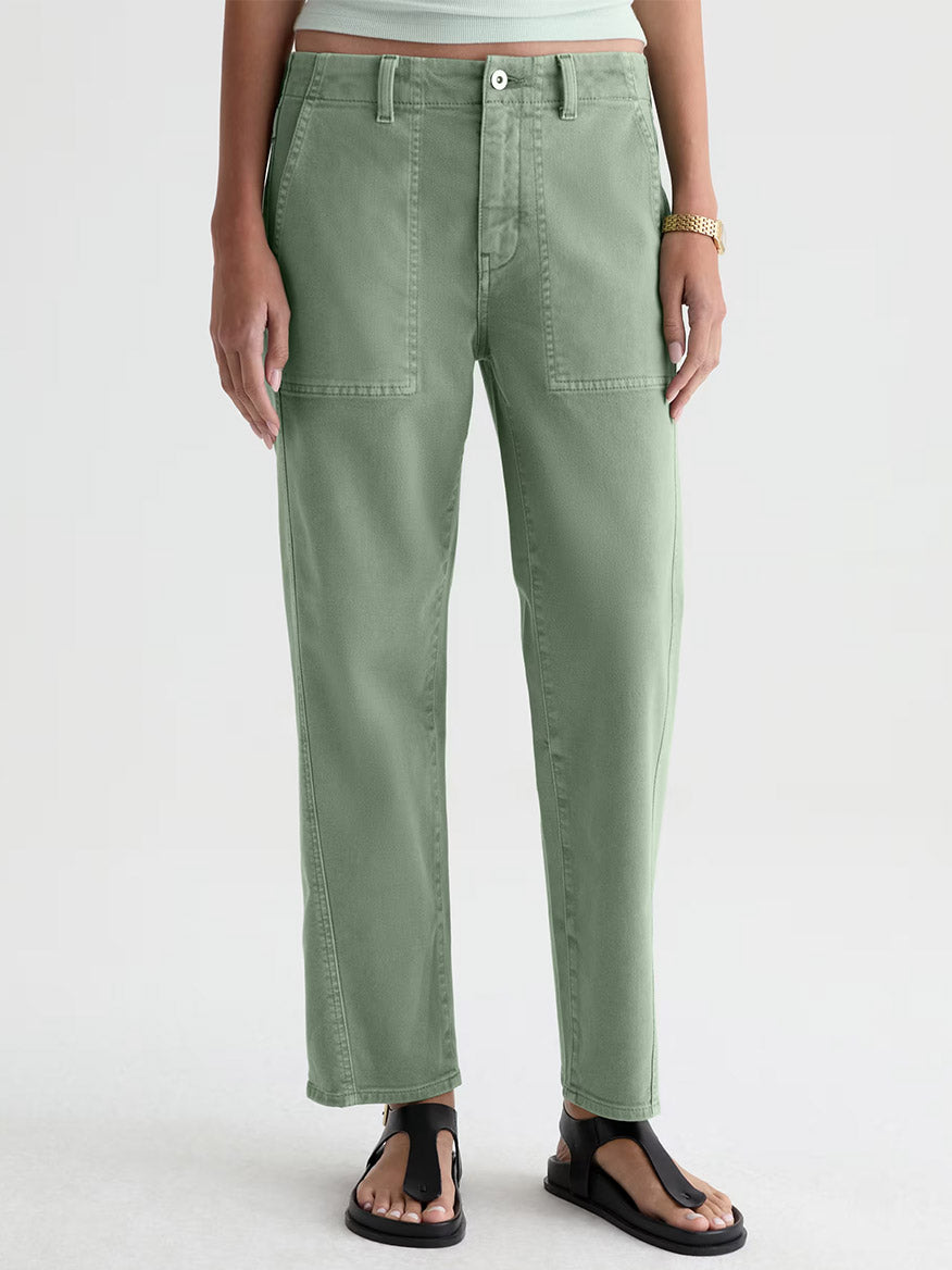 Someone wearing AG Jeans Analeigh Mid-Rise Utility Pant in Sulfur Faded Moss, a white shirt, gold watch, and black sandals stands against a plain light background.
