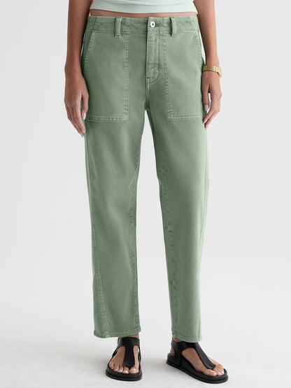 Someone wearing AG Jeans Analeigh Mid-Rise Utility Pant in Sulfur Faded Moss, a white shirt, gold watch, and black sandals stands against a plain light background.