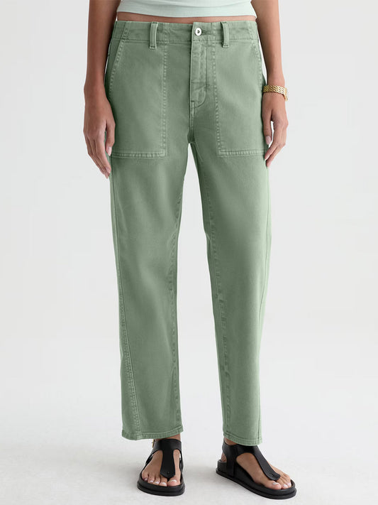 Someone wearing AG Jeans Analeigh Mid-Rise Utility Pant in Sulfur Faded Moss, a white shirt, gold watch, and black sandals stands against a plain light background.