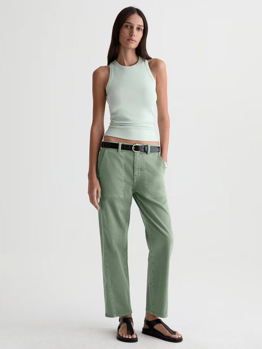 A woman stands against a plain background wearing a light green sleeveless top, AG Jeans Analeigh Mid-Rise Utility Pant in Sulfur Faded Moss with a black belt, and black sandals.