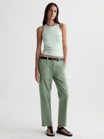 A woman stands against a plain background wearing a light green sleeveless top, AG Jeans Analeigh Mid-Rise Utility Pant in Sulfur Faded Moss with a black belt, and black sandals.