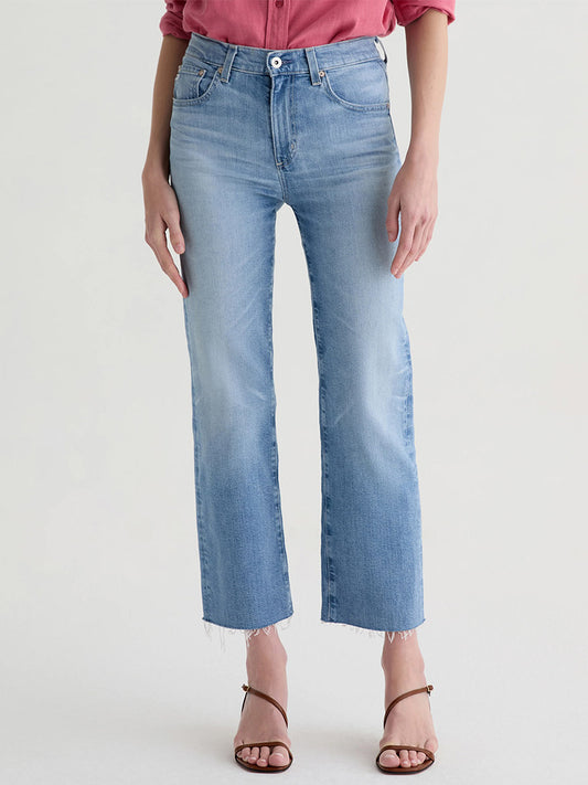 A person wears AG Jeans Brinley Mid-Rise Straight in Fiction—light blue, vintage comfort stretch denim with raw hems—paired with a pink long-sleeve shirt and brown strappy sandals, standing against a plain background.