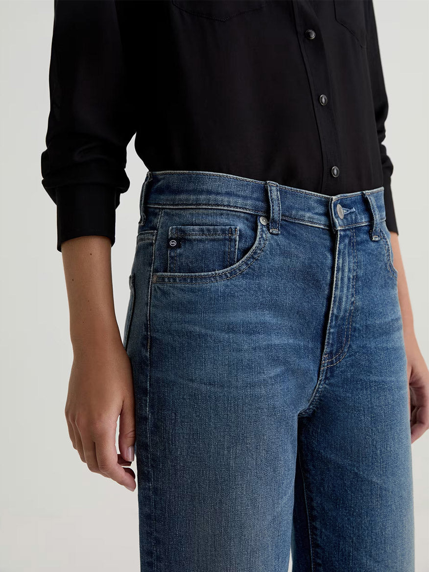 Model wearing the AG Jeans Brinley Mid-Rise Straight in Hotel California, a high-waisted blue jean in comfort stretch denim, paired with a black long-sleeve button-up shirt; shown from mid-torso to upper thigh.
